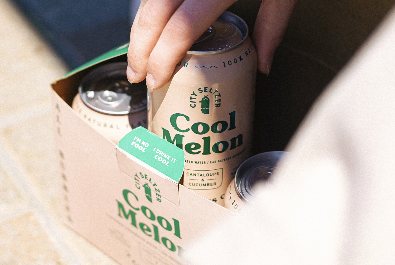 Hand pulling a can of Cool Melon seltzer from the cool melon-coloured case.